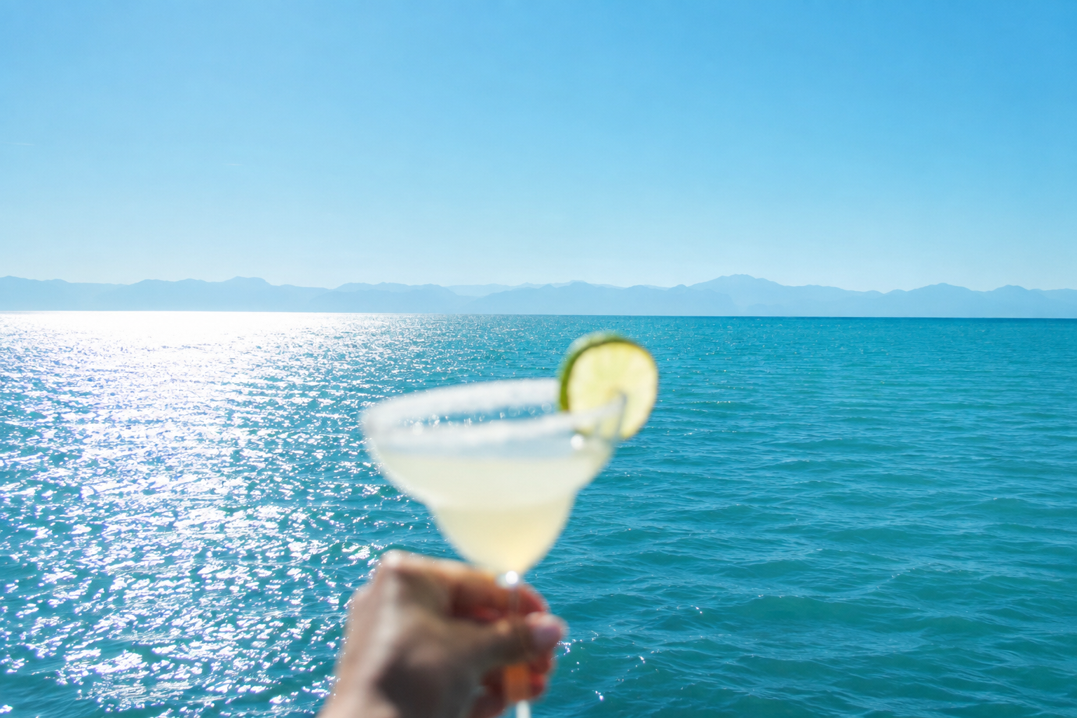 Hand holding a salt-rimmed margarita against a bright blue sea and sky